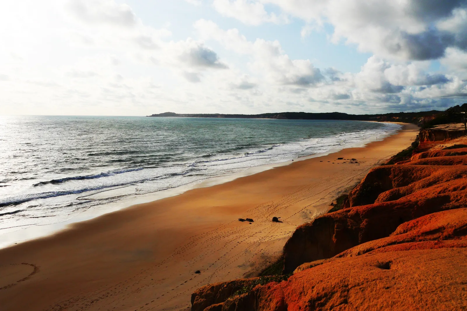 A long beach at Cacimbinhas seen from the top of the red cliffs, footprints across the sand, waves running in from the left and a low wooded headland on the horizon