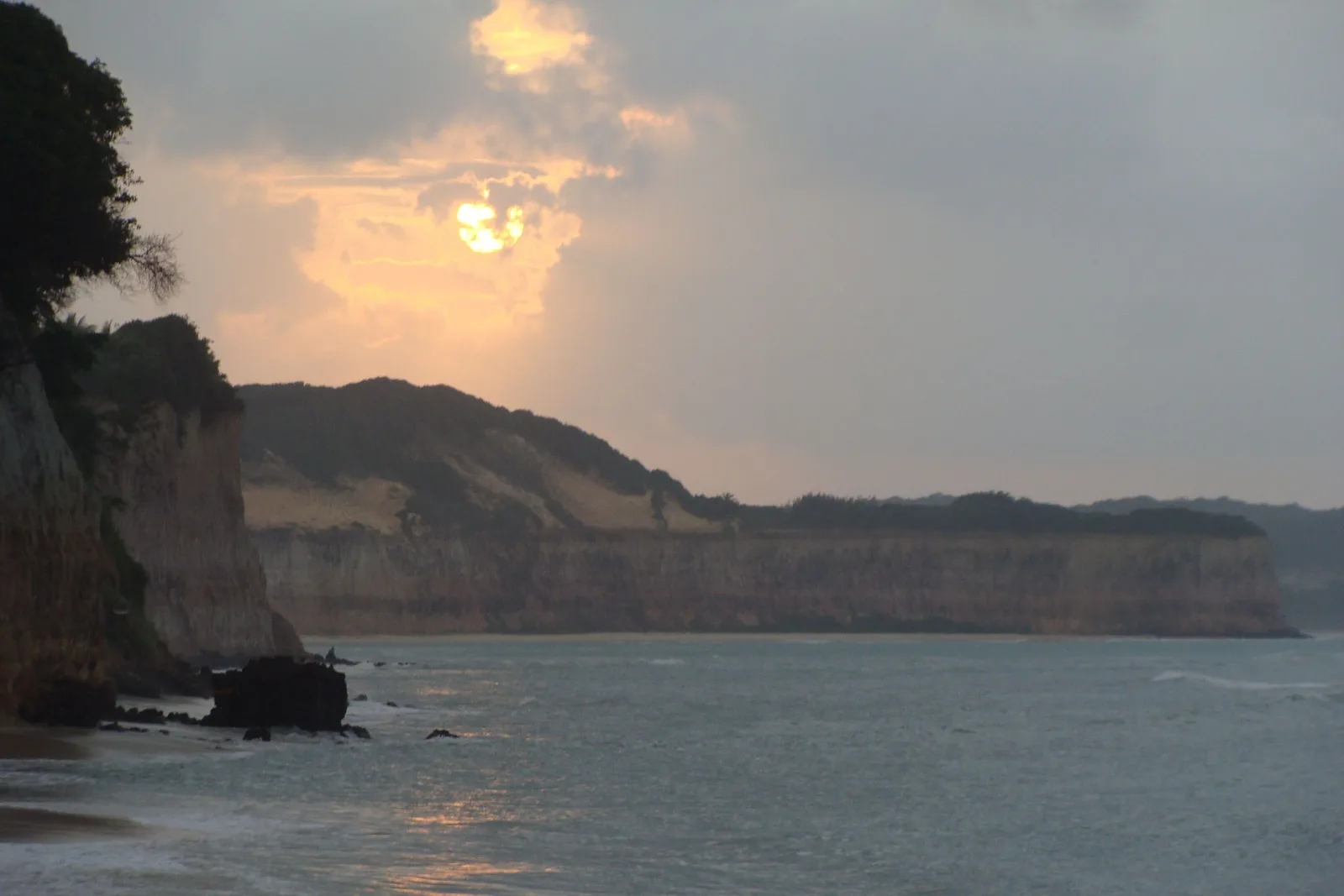 The sun low behind cloud above the cliff wall at Pipa, seen along the beach with dark rock standing in the shallows and the pale cliff line running away into haze