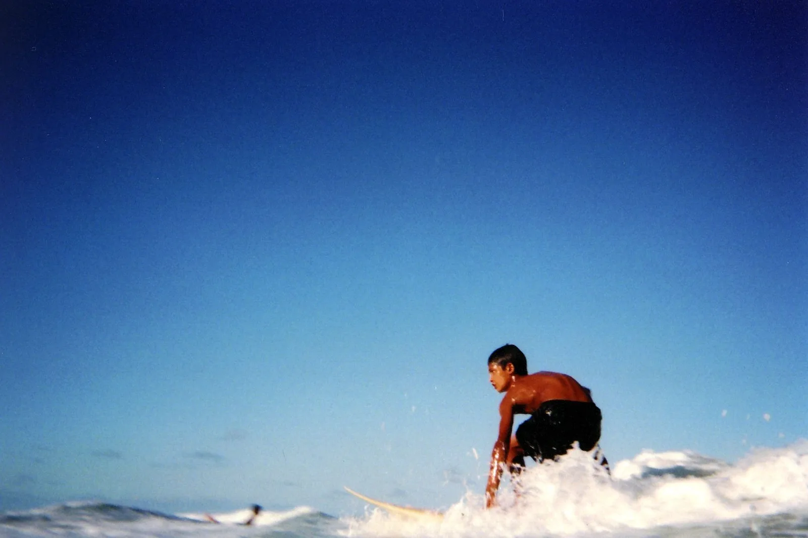 A young surfer crouched low on a board in the white water of a breaking wave, seen side-on from water level against a deep blue sky, with a second surfer lying on a board at the left