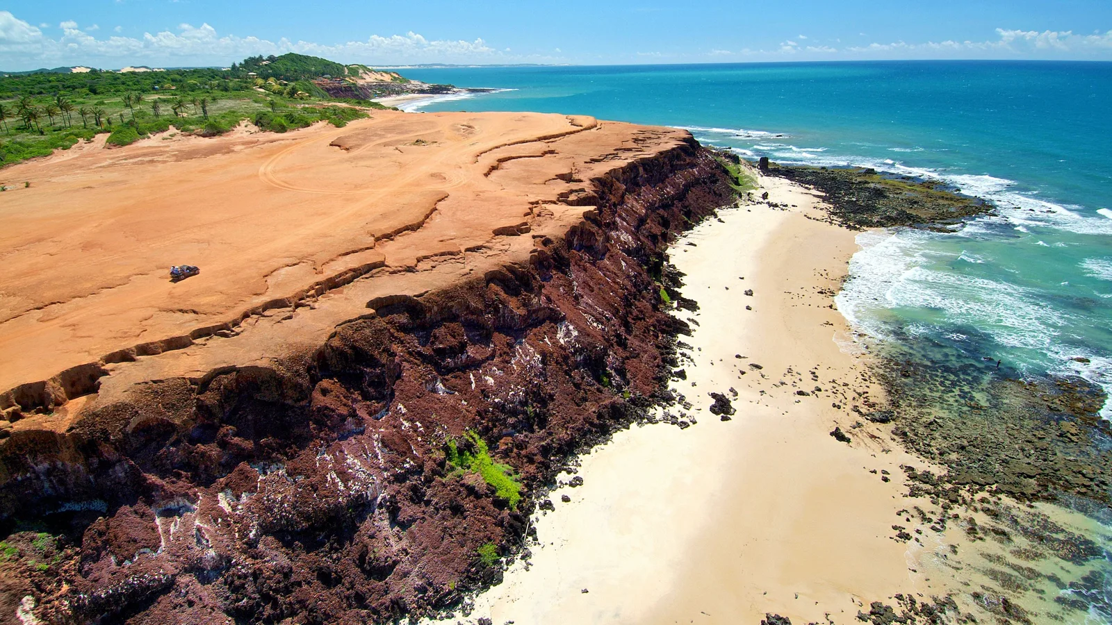 The flat red top of the Chapadão from the air with a vehicle parked on it, its dark cliff face dropping to a strip of white sand, and rock ledges and turquoise surf beyond
