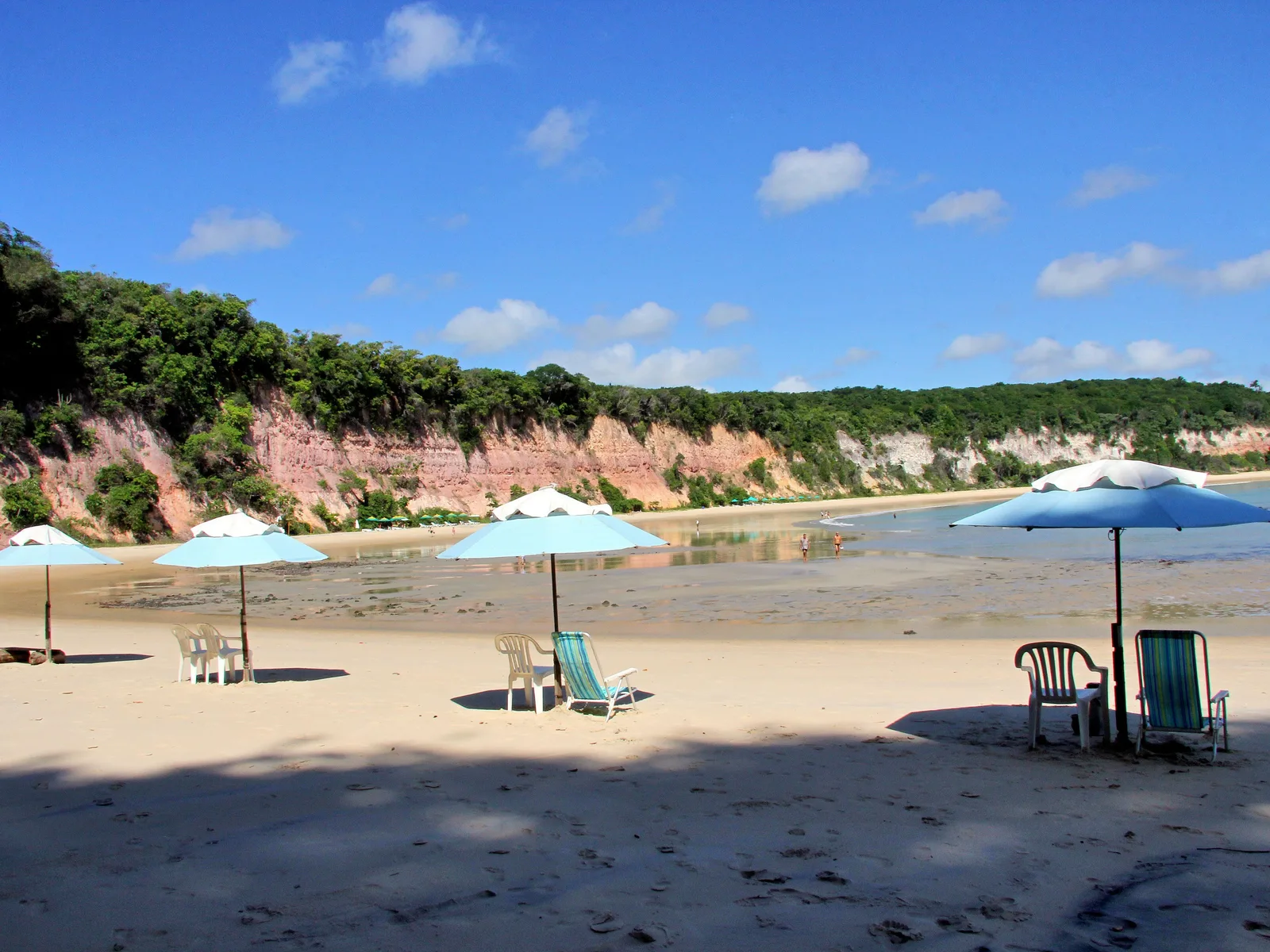Blue and white parasols and empty beach chairs on the sand at low tide below a long wooded red cliff at Pipa, a few people far off at the water’s edge
