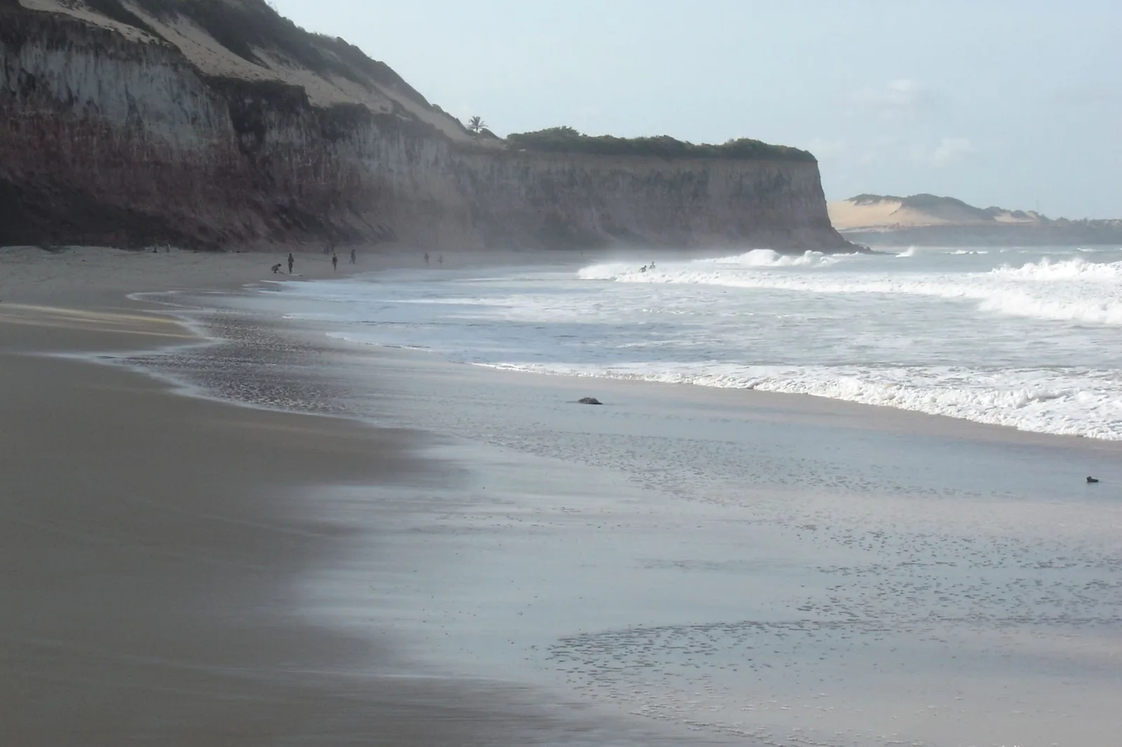 A wide stretch of wet sand exposed at low water below the cliffs at Pipa, with a line of surf beyond it and people small in the distance