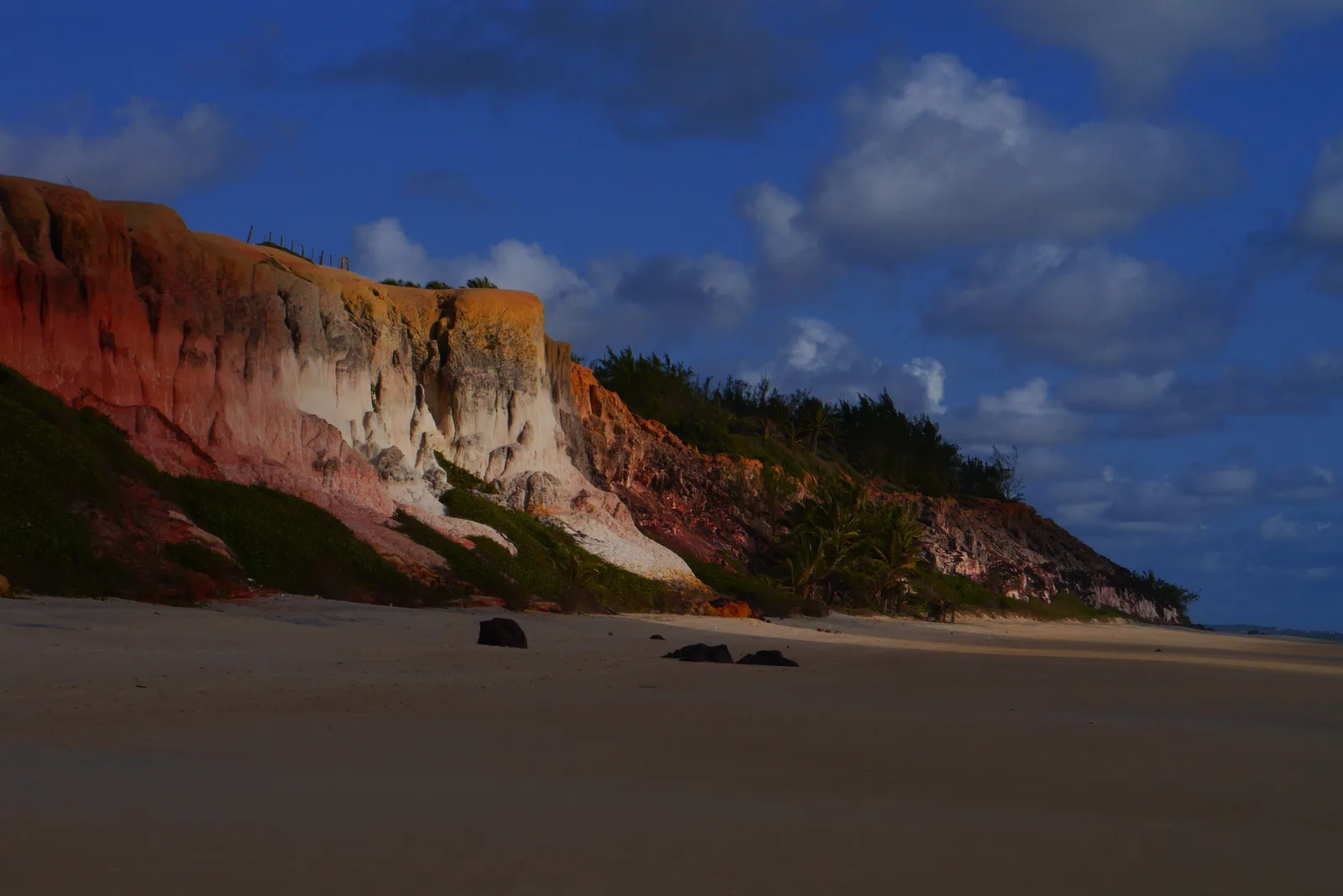 A banded red and cream cliff face rising straight off the sand at Praia de Cacimbinhas under a blue sky with heavy white cloud