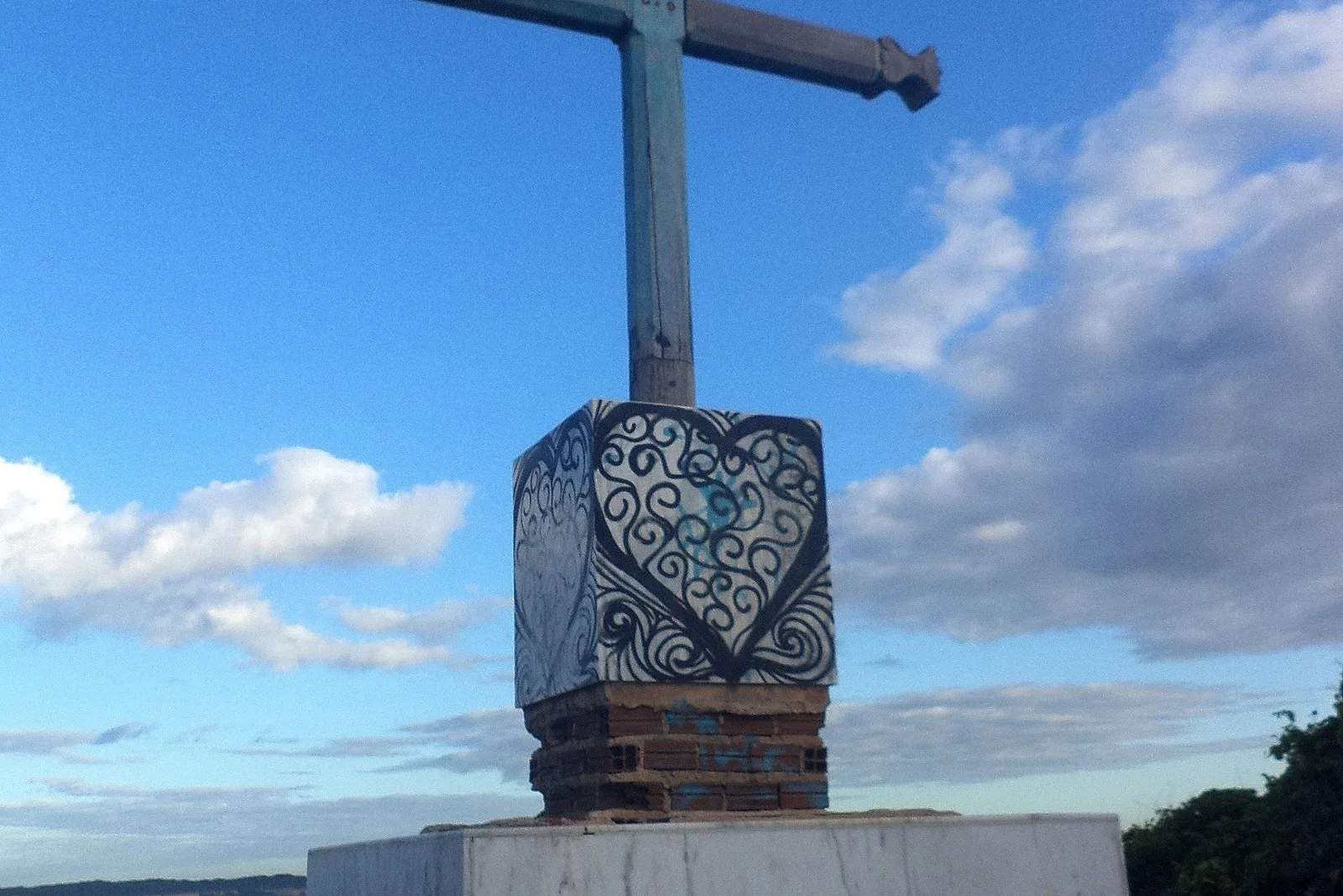 A metal cross in Pipa mounted on a plastered pedestal painted with a black and white heart and scroll design, standing against a blue sky with scattered white cloud