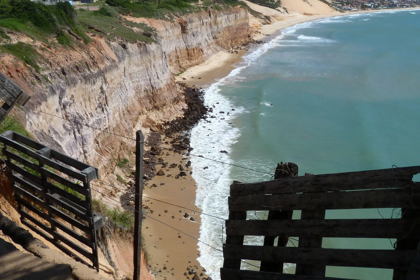 Looking down a long eroded cliff wall on the Rio Grande do Norte coast past a rough wooden gate and fence, a narrow strip of beach and dark rock at its foot, turquoise water along the shore and a town and dunes in the distance