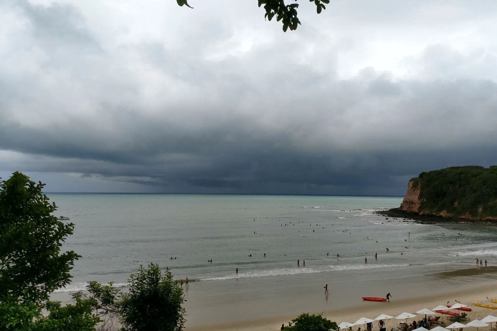 Heavy storm cloud sitting over the bay at Praia do Madeiro, swimmers and surfers spread across gray-green water and parasols and kayaks along the sand below