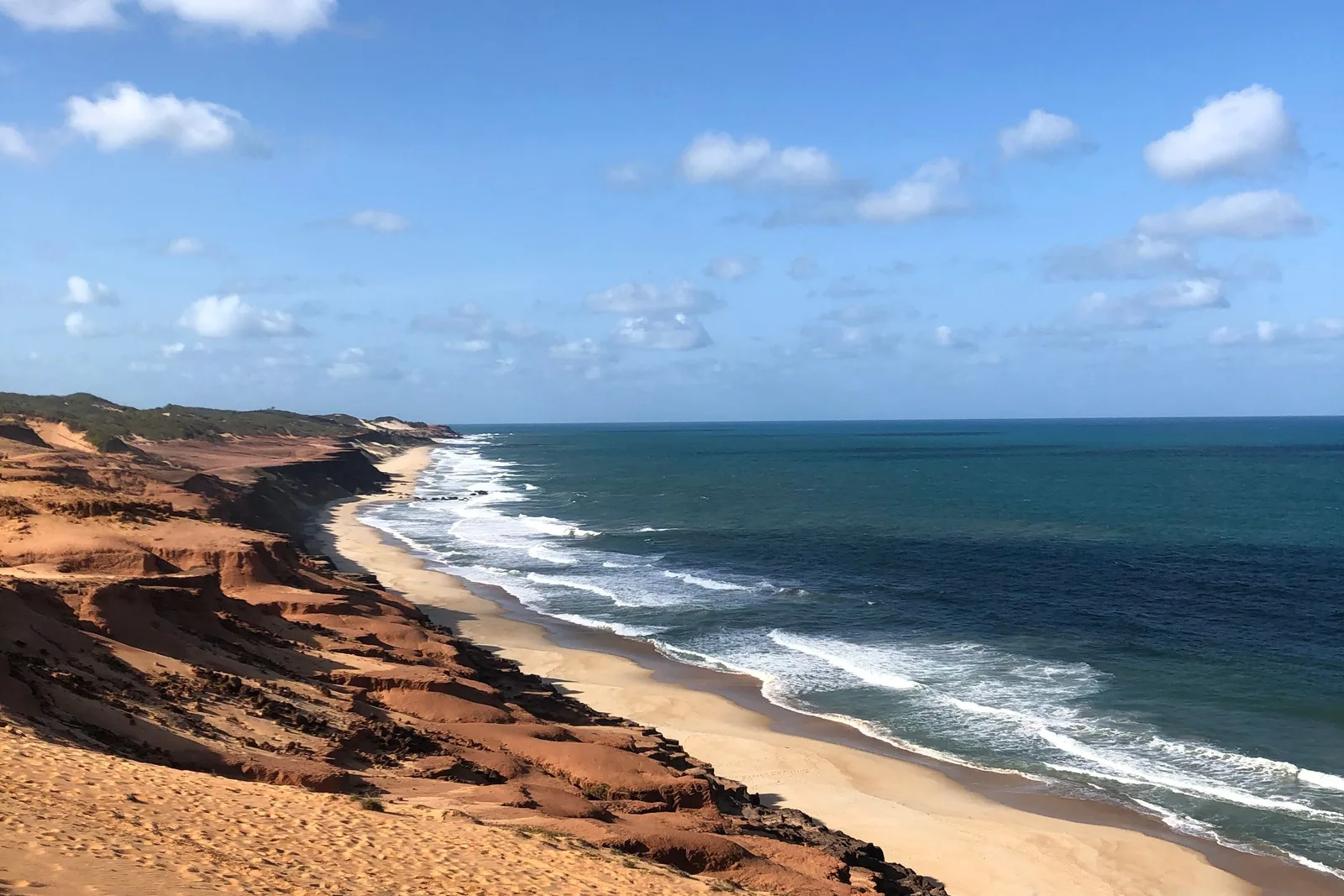 A long empty beach seen from a red clifftop, the eroded cliff line and pale dunes running away to the north and open blue ocean filling the right of the frame