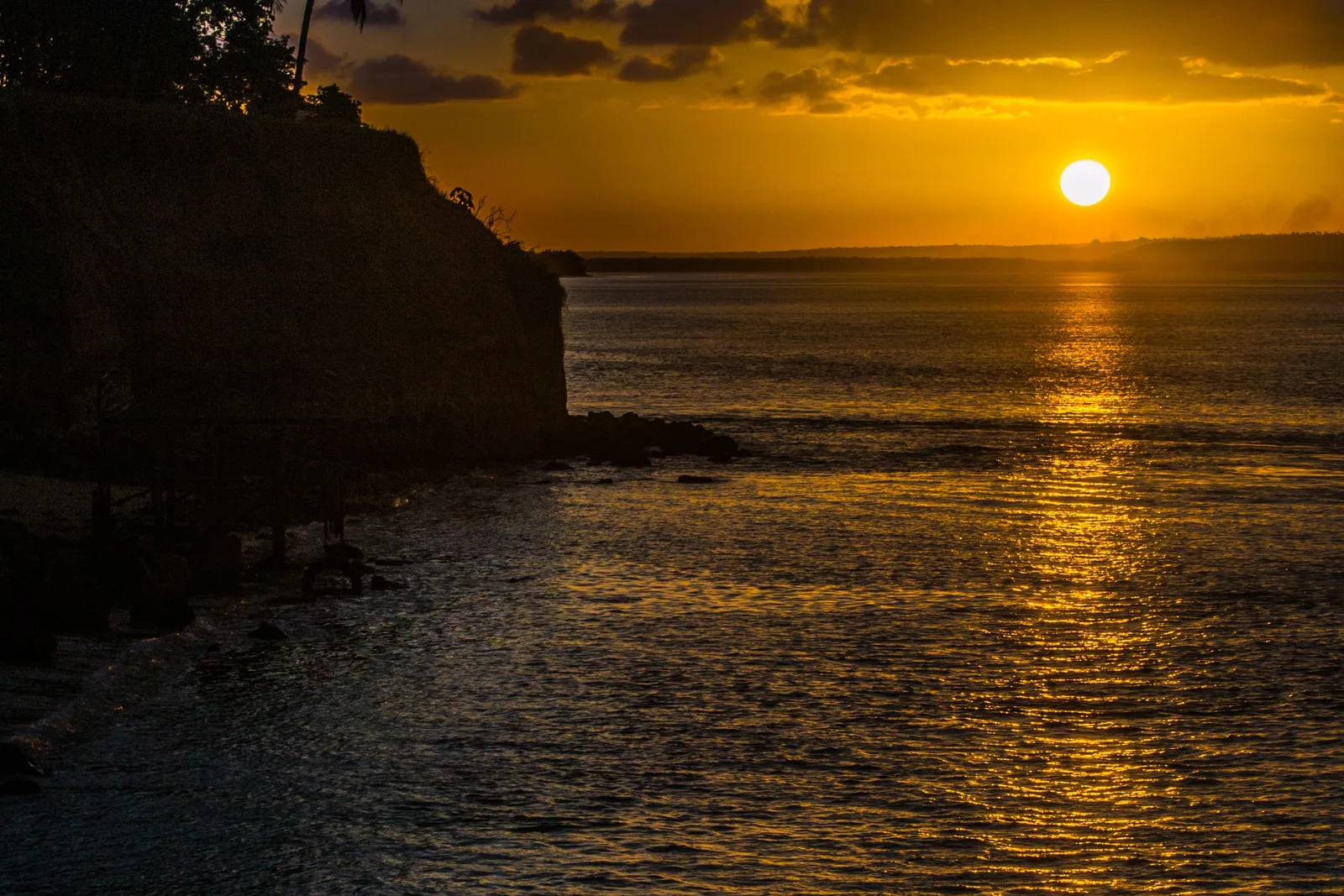 A gold sunset over calm water on the Tibau do Sul coast, a dark cliff, palms and a jetty in silhouette on the left and a low far shore along the horizon