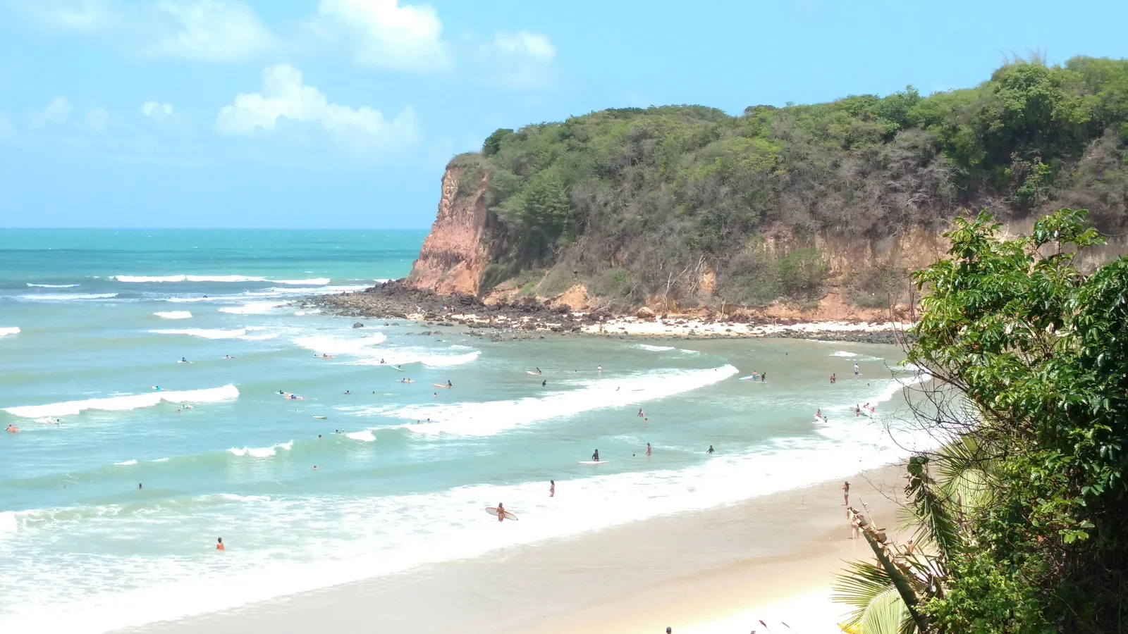 A green-topped headland with a bare red cliff face closing the far end of a beach at Pipa, dozens of surfers and swimmers spread across the turquoise water and a wide curve of pale sand in the foreground
