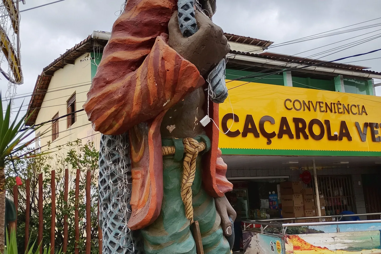 The painted body of a large fisherman sculpture in Pipa, its head out of the frame, a net slung over its shoulder and a rope for a belt, in front of a convenience store’s yellow sign