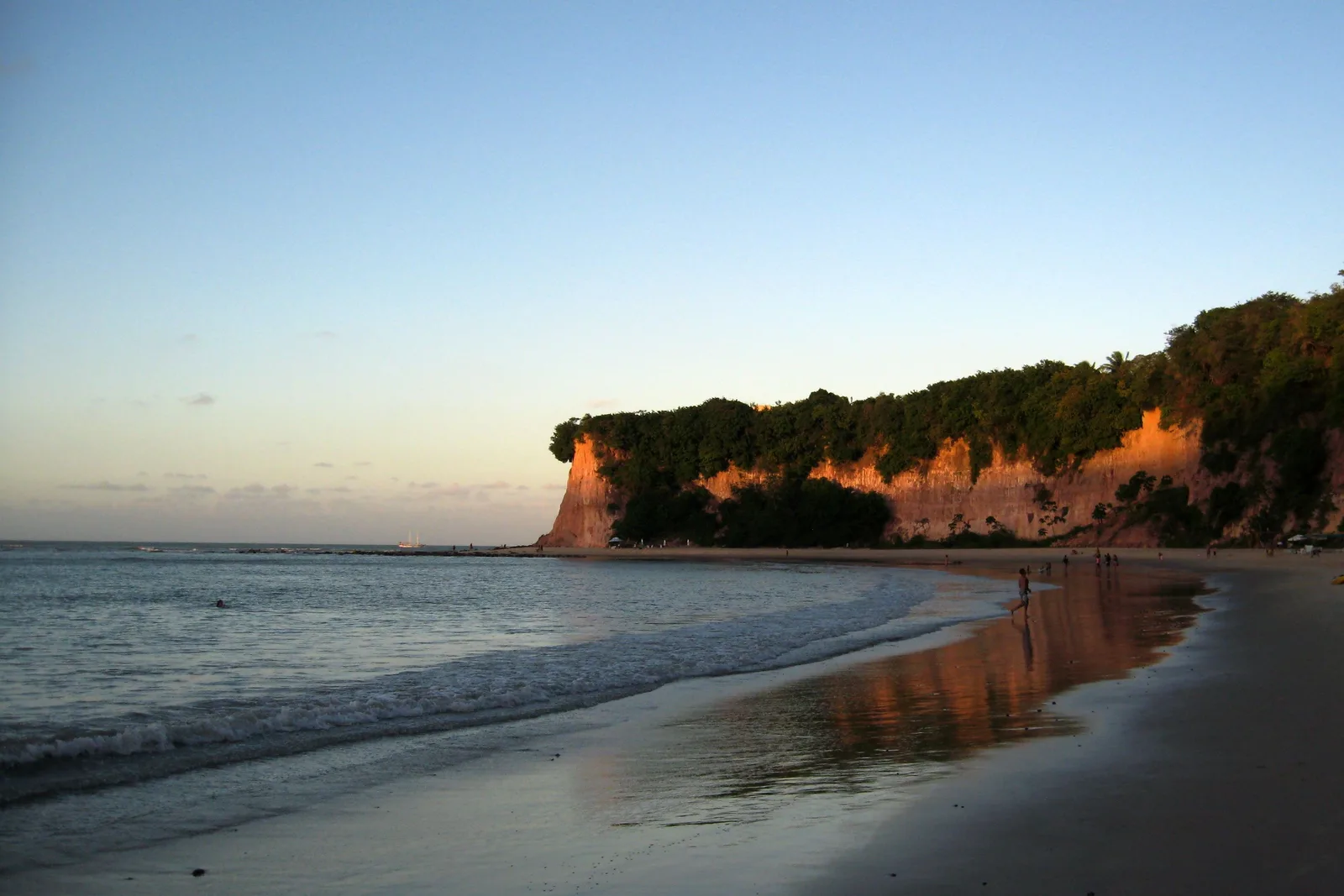 A wooded red cliff headland lit orange by low sun at Pipa, reflected in the wet sand of the beach below, with a few people at the water’s edge and small waves running in