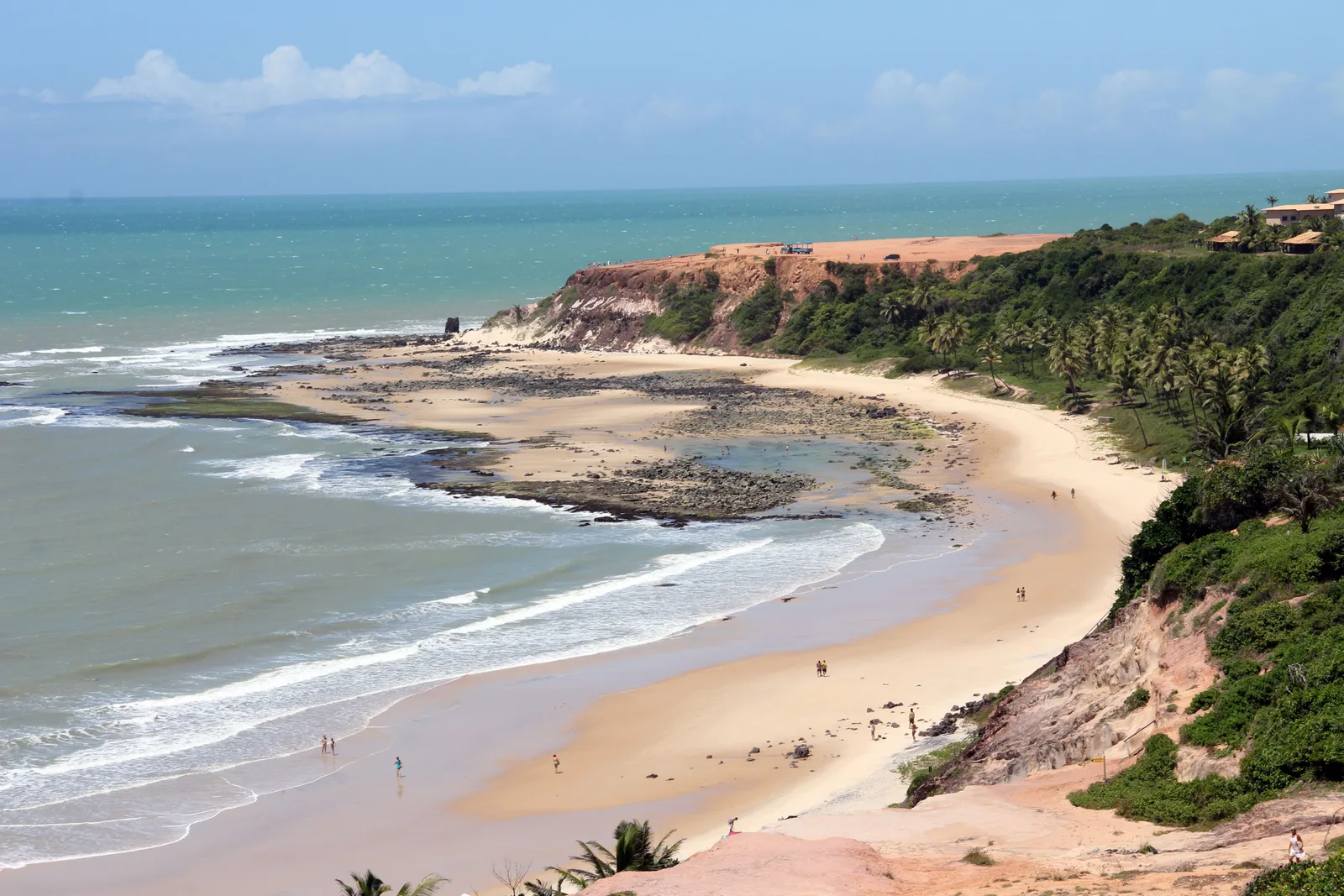 Praia do Amor seen from above: a curving beach below palm-covered slopes, wide dark rock ledges exposed in the shallows, a few people on the sand and a flat-topped red headland at the far end