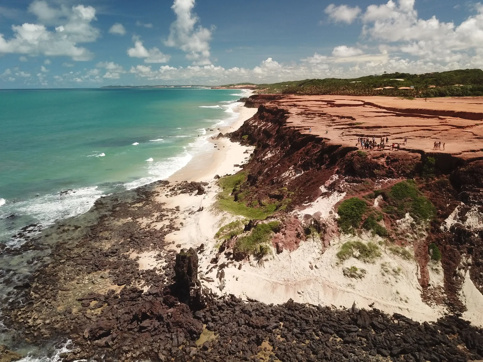 The Chapadão seen from the air: a flat red rock plateau with a group of people standing at its edge, dark rock at its base, and a pale beach and turquoise sea running away along the coast behind it