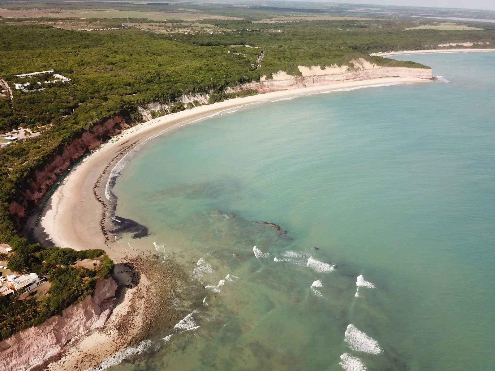 Baía dos Golfinhos from the air: a wide curve of pale sand under forested red and white cliffs, shallow turquoise water across the bay and small waves breaking over dark rock near the headland in the foreground