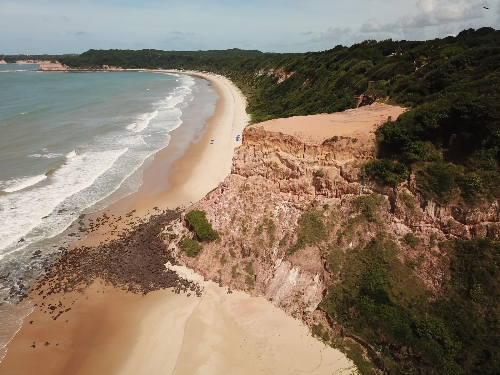 Praia do Madeiro from the air: a bare, banded red headland with dark rock at its foot in the foreground, and a long curve of pale sand running away under wooded cliffs with lines of surf breaking along it