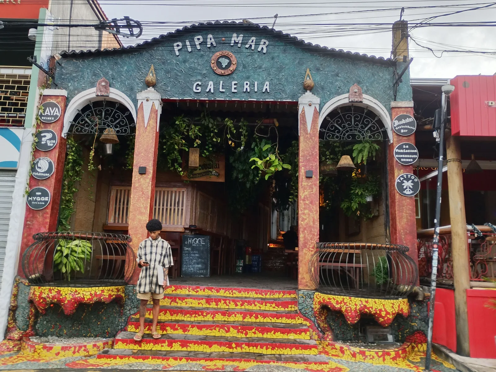 The front of a small shopping arcade in Pipa with painted arches, hanging plants inside and mosaic-tiled steps, and a young man standing on the steps looking at his phone