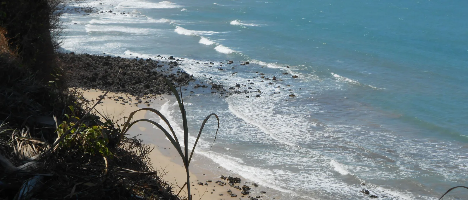Looking straight down from the clifftop at Praia do Amor onto a narrow strip of sand scattered with dark rock, lines of surf running in over blue water