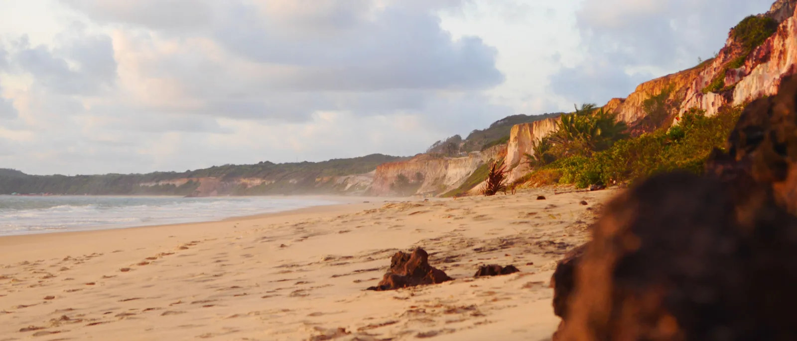A wide sandy beach in warm low light at Cacimbinhas, dark rock in the foreground and a red and cream cliff wall running away along the shore under green vegetation
