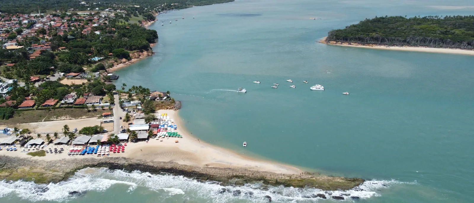The town of Tibau do Sul from the air where the Guaraíras lagoon meets the sea, with boats on the pale water, a sand spit between lagoon and ocean and the town along the shore