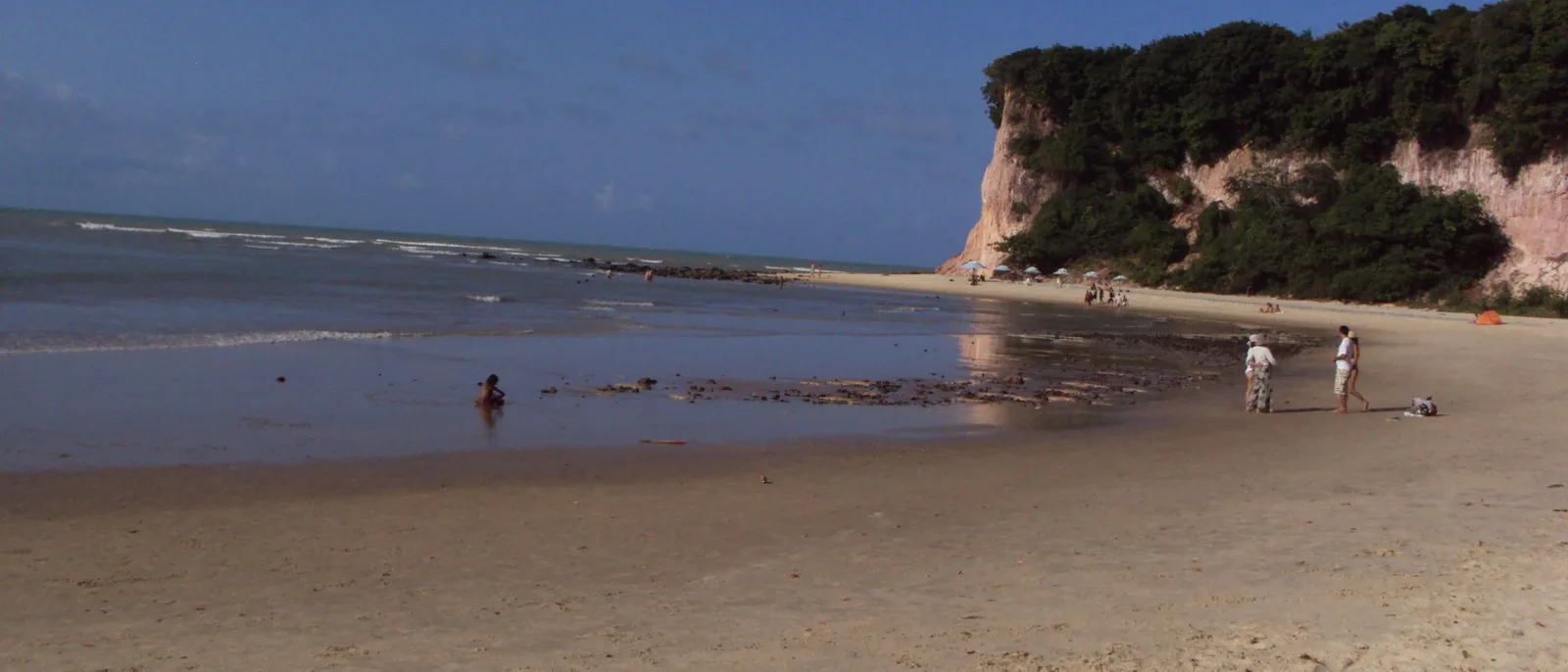 The wide low-tide beach in front of Pipa with wet sand in the foreground, white cliffs closing the far end of the bay and a few people walking along the water’s edge