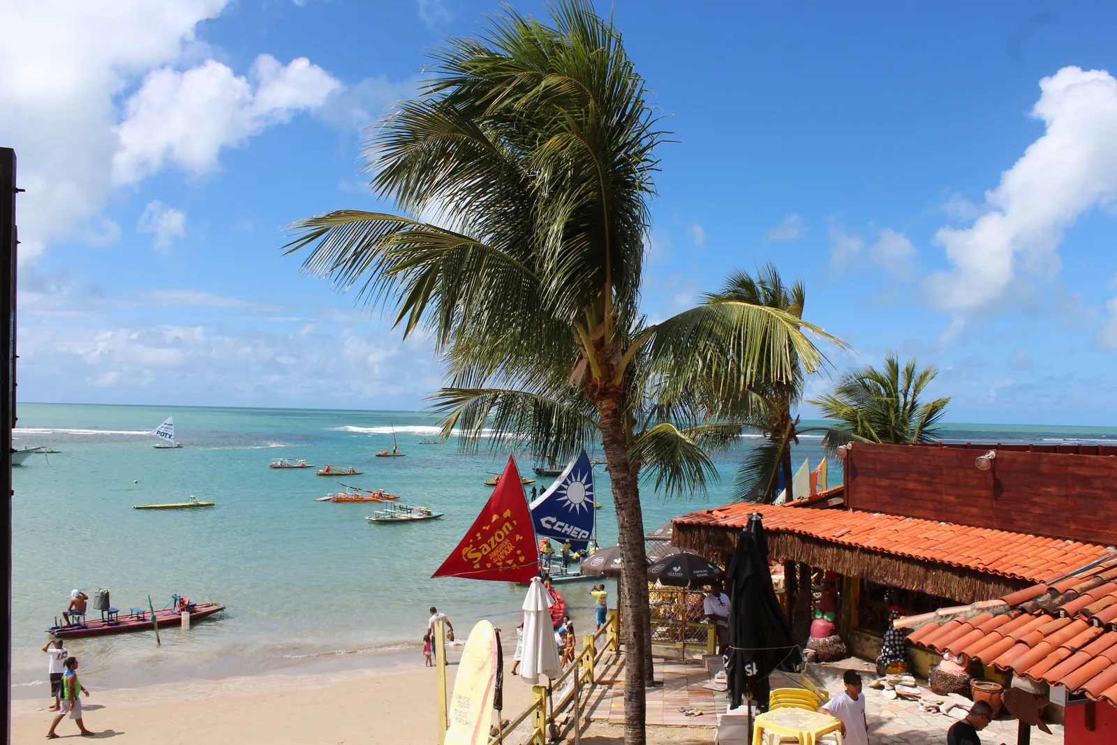 A red-roofed beach bar under coconut palms at Porto de Galinhas with sun umbrellas around it and jangadas drawn up on the turquoise water beyond