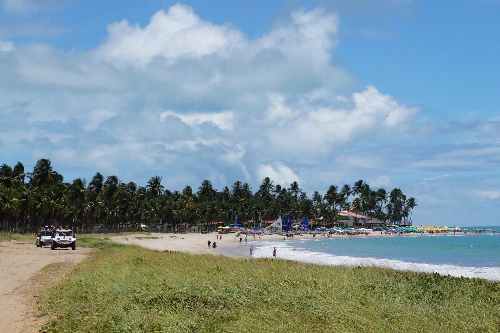 Two white dune buggies with passengers parked on a sand track behind the beach at Porto de Galinhas, a long palm-backed shore with kiosks and umbrellas stretching away beyond them