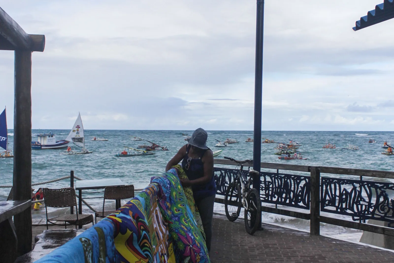 A vendor leaning on a seafront railing at Porto de Galinhas beside brightly printed cloths hung out for sale, with boats on the water behind