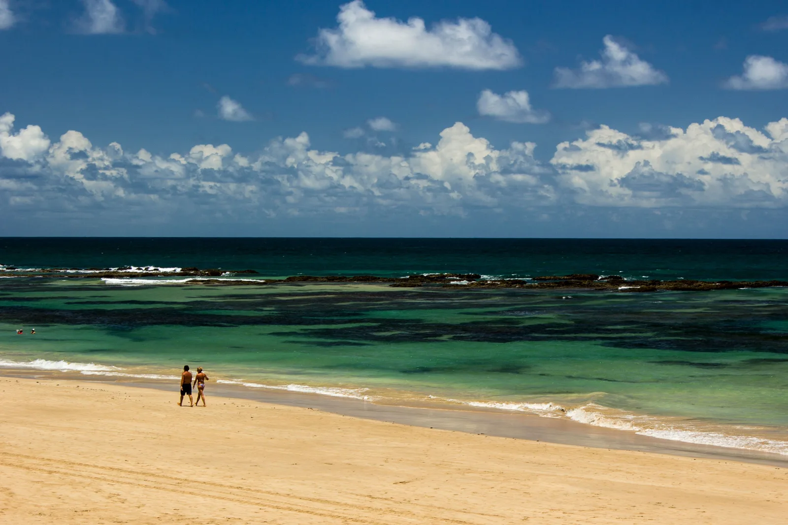 A wide open beach at Cupe with two figures walking on the wet sand and a band of green reef water offshore under a cloudy sky