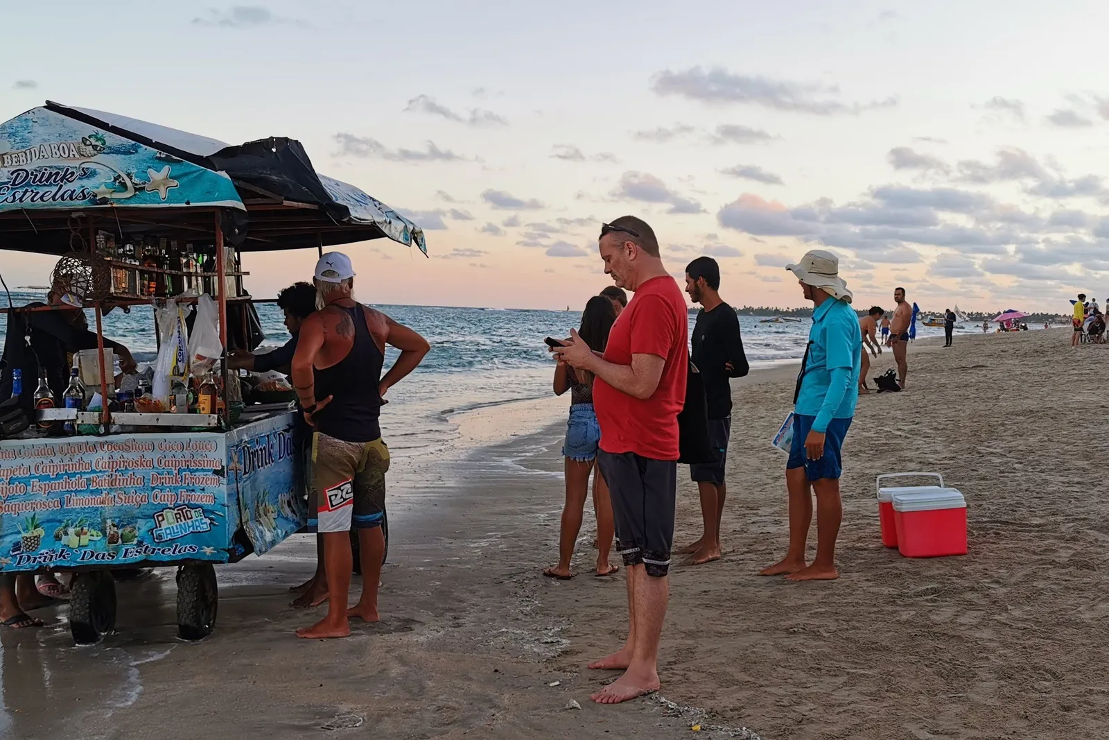 A brightly painted drinks cart on the sand at Porto de Galinhas at dusk, a vendor serving a small group of customers beside it
