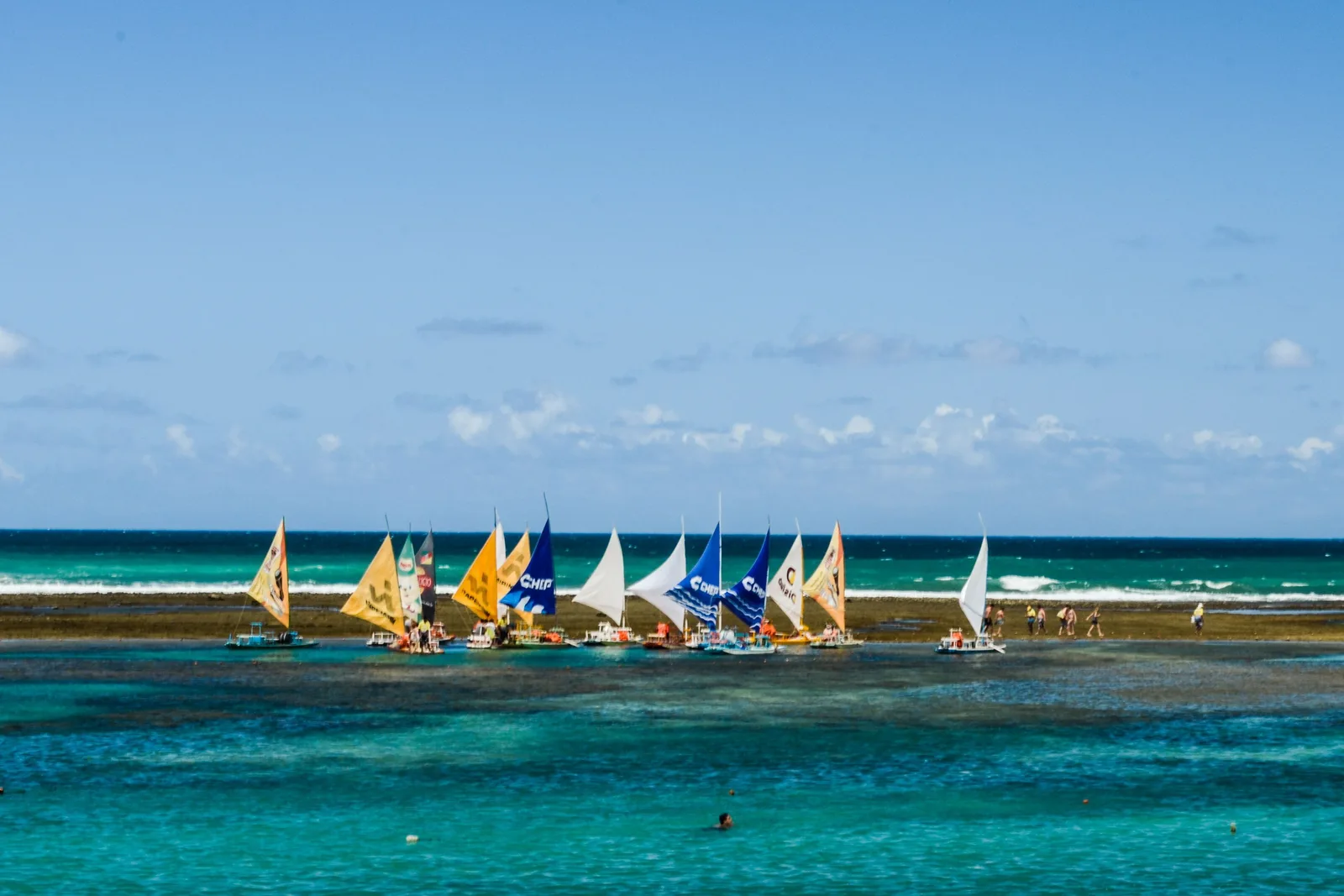 A line of jangadas with striped red, blue and yellow sails anchored along the reef at Porto de Galinhas in bright turquoise water, people standing in the shallows around them
