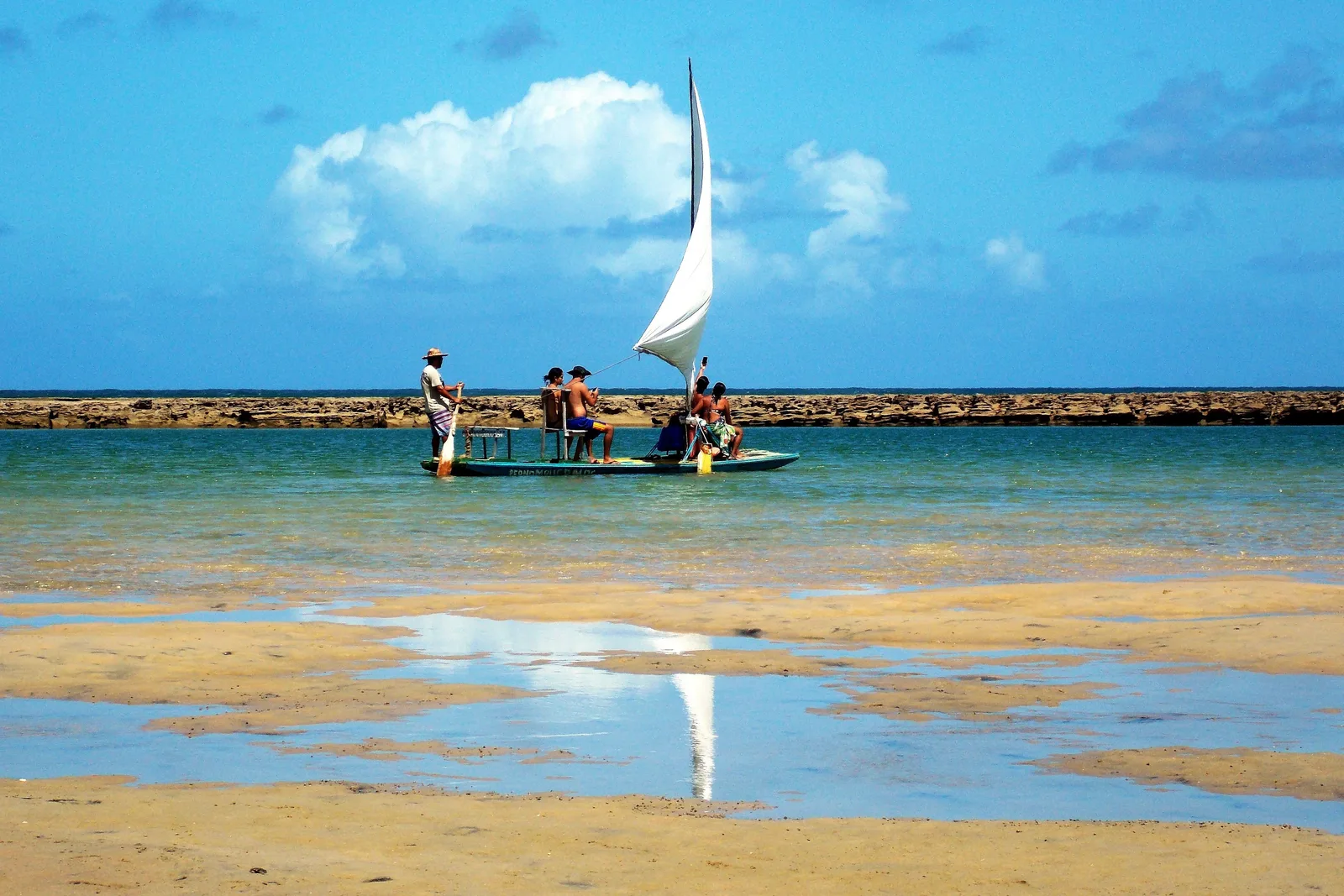 A jangada with a white sail resting on a wet reef flat at Muro Alto, people standing beside it and their reflections in the mirror-still shallow water