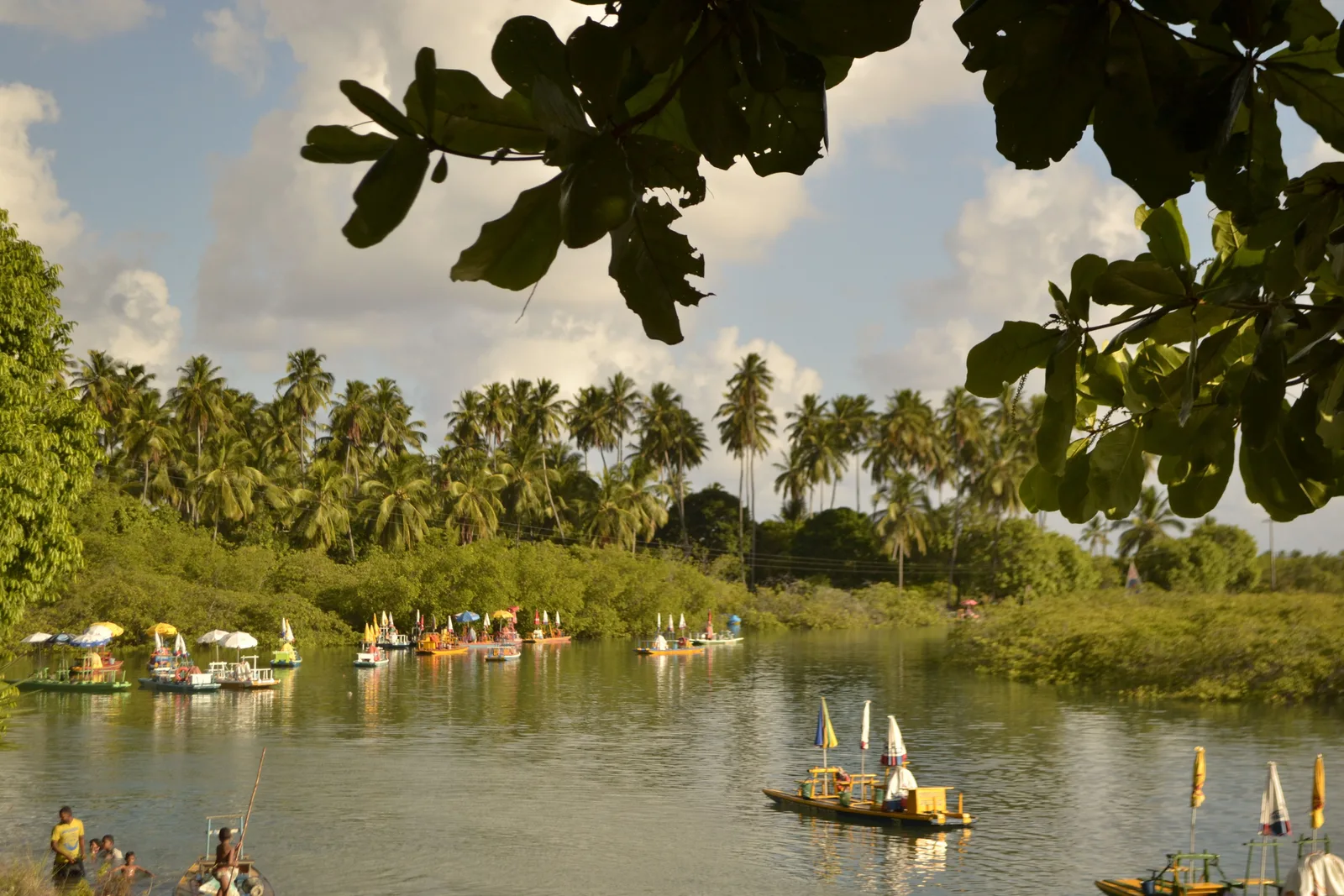 The shallow estuary at Pontal de Maracaípe seen from under overhanging branches, jangadas and small boats drawn up along the far bank with people wading in the water