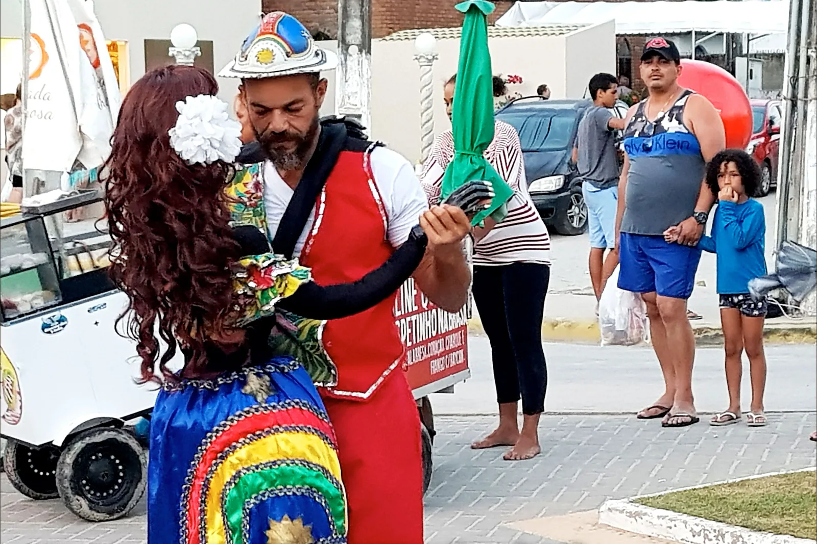 A costumed street performer in a wig and a red and white outfit holding a green flag on a street in Porto de Galinhas, a family and an ice cream cart beside them