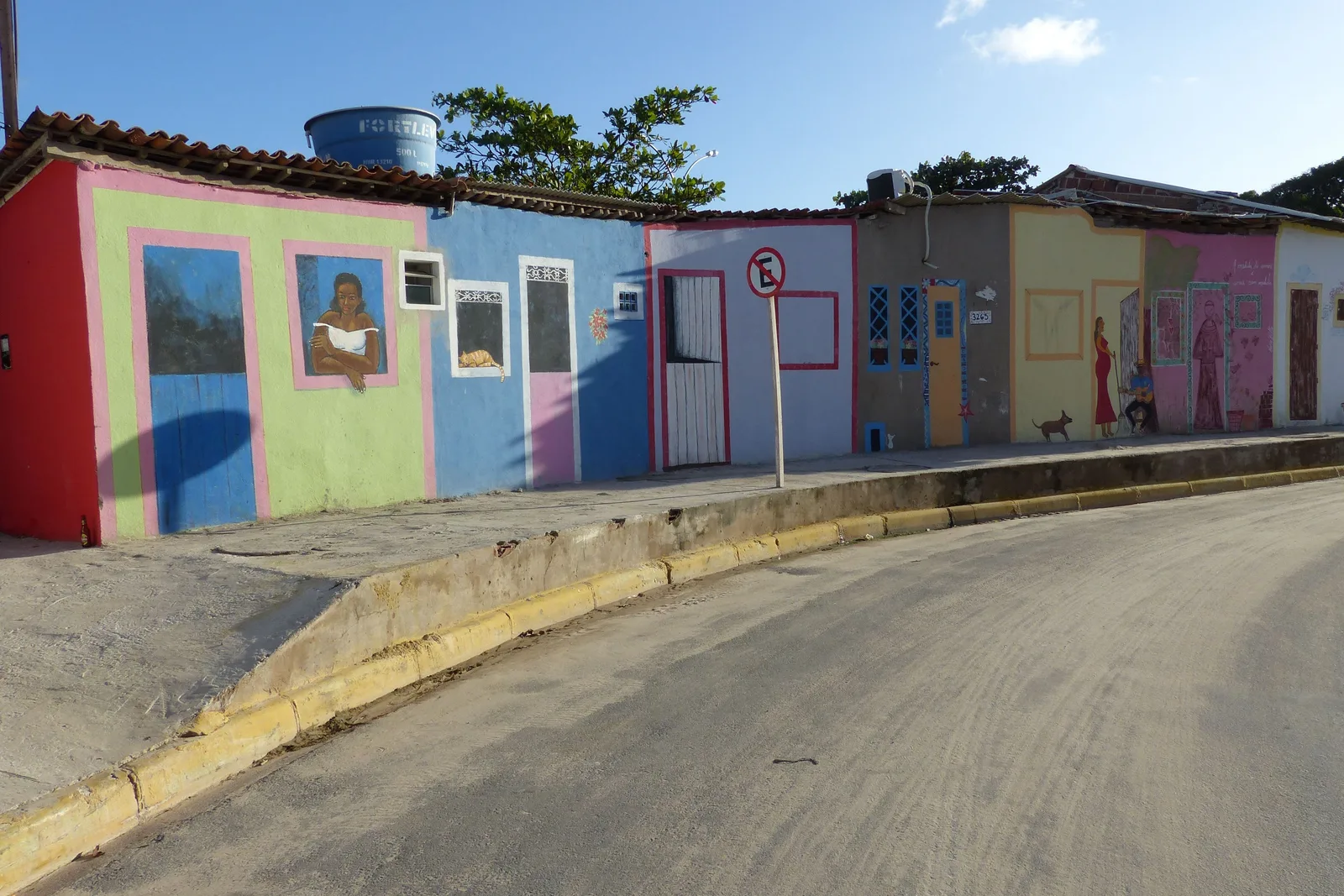 A quiet street in Porto de Galinhas lined with small houses painted in flat pink, blue, green and yellow, a curved road running past them