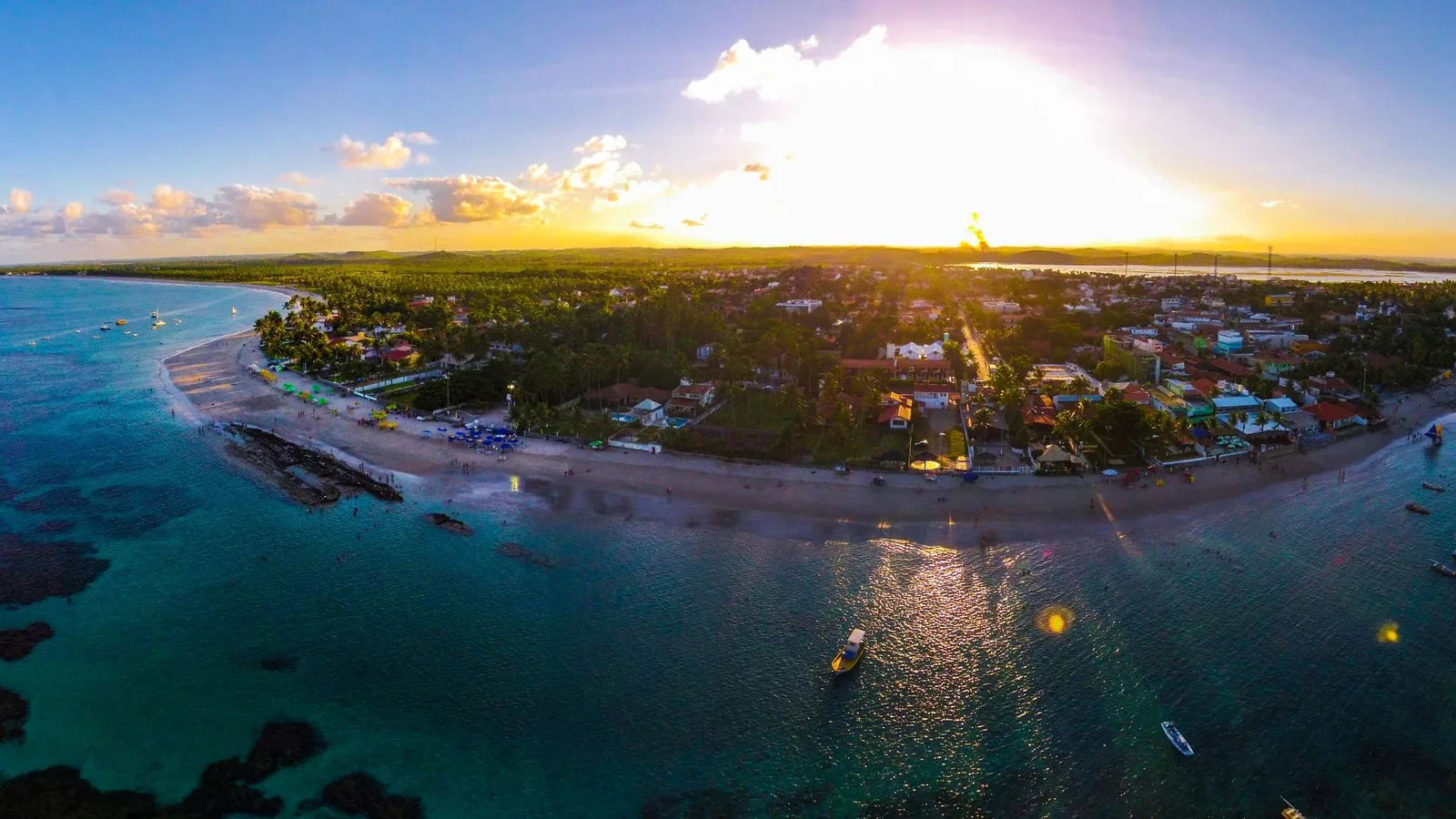 Porto de Galinhas from the air at sunset, the village lights coming on along the shore and the reef line dark in the pale water offshore