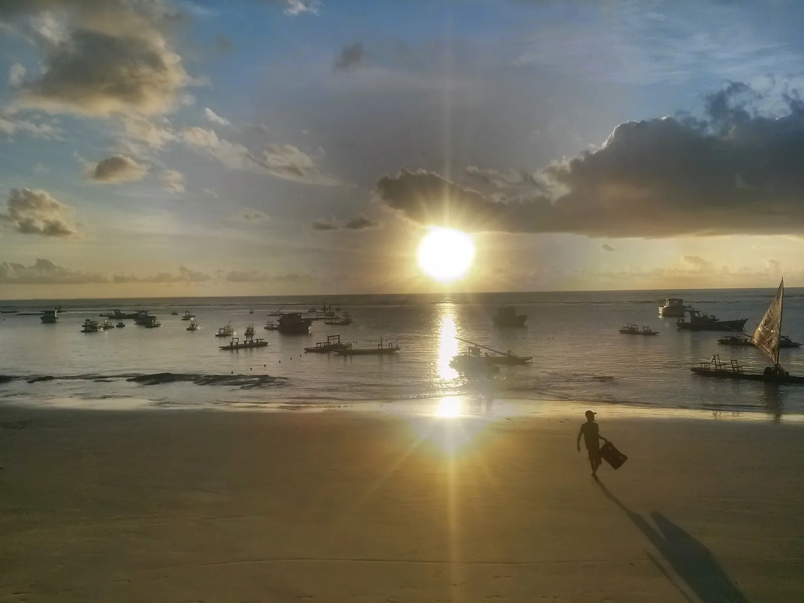 Sunrise over broad reef flats at Porto de Galinhas exposed by a very low tide, boats moored beyond and a figure walking on the wet rock