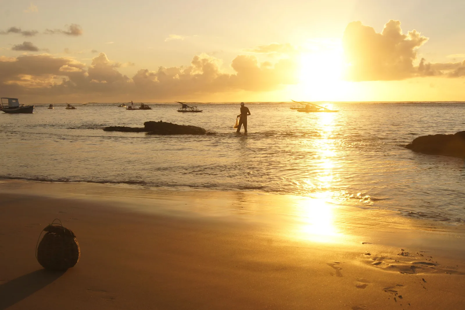 A figure wading in shallow water at dawn at Porto de Galinhas, the low sun laying a band of gold across the wet sand and moored boats behind
