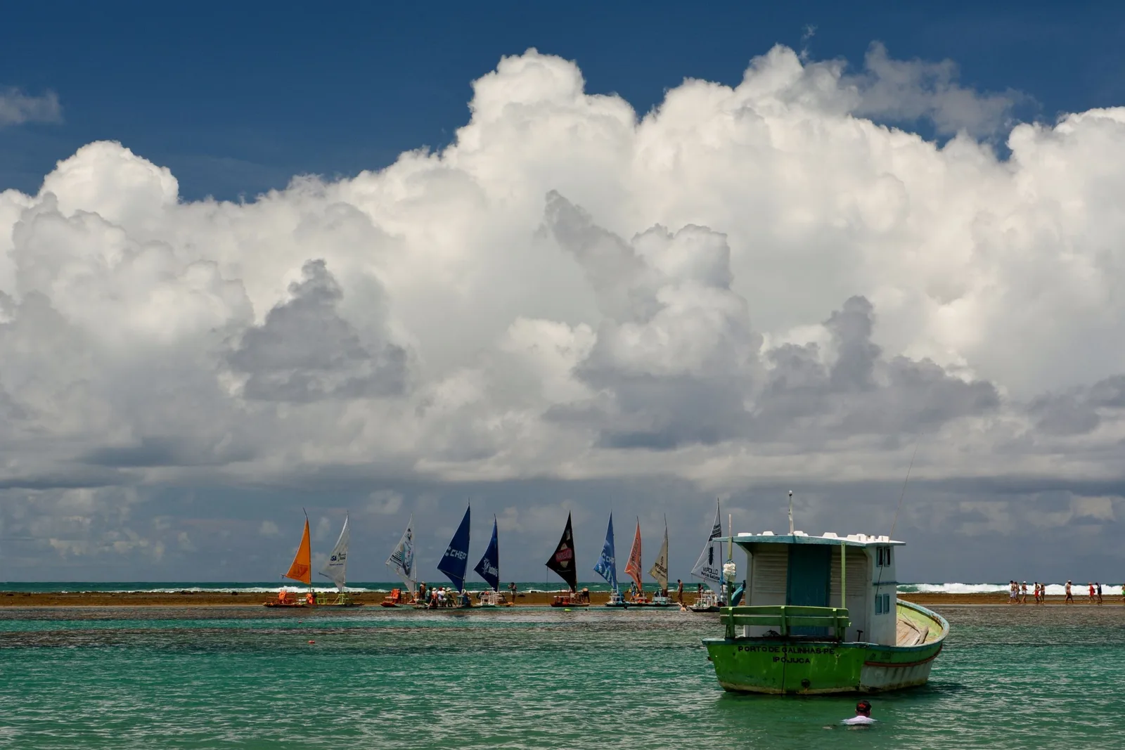 A line of jangadas with colored sails moored along the reef at Porto de Galinhas, a small green launch beside them and towering white cloud above