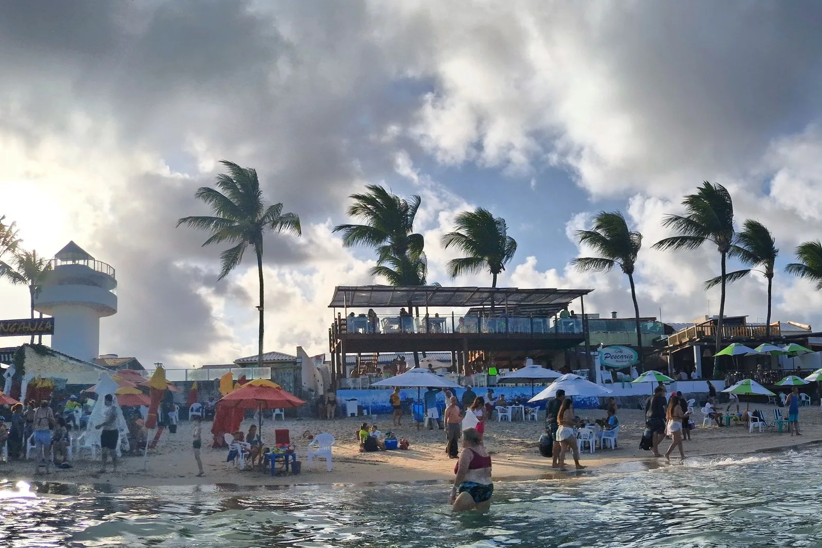 The main beach at Porto de Galinhas on a busy day: a crowd under sun umbrellas along the sand, kiosks and coconut palms behind them and low gray cloud overhead