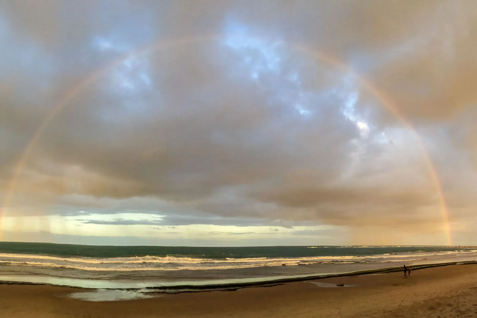 A faint rainbow standing over the surf on a wide beach at Porto de Galinhas under heavy cloud