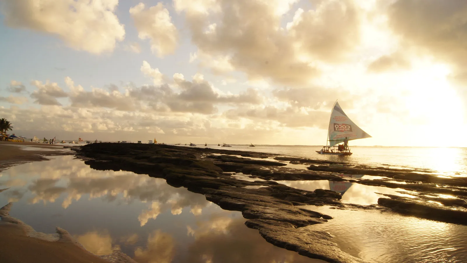 A jangada with its sail up resting on a long spit of exposed reef at sunrise, the wet rock and shallow water either side of it mirroring a gold and gray sky