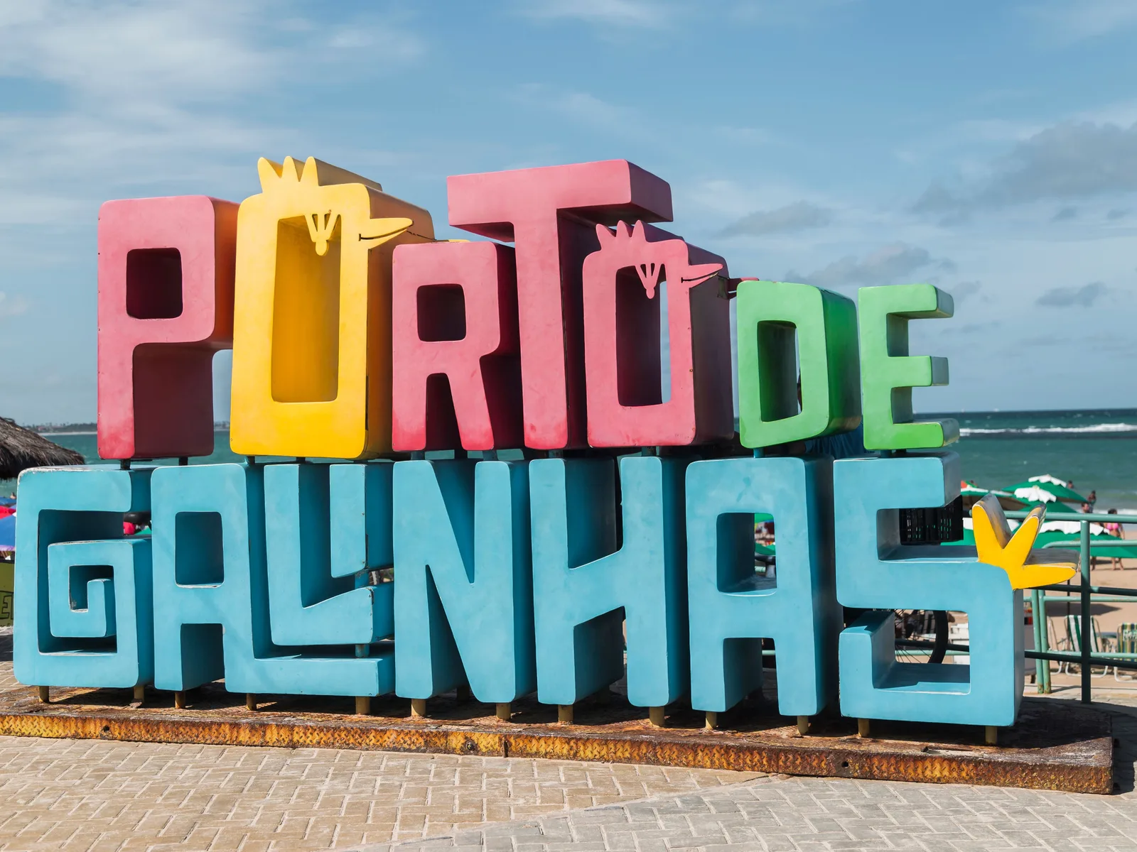 The tall painted letters spelling PORTO DE GALINHAS standing on the seafront, with people and beach umbrellas behind them