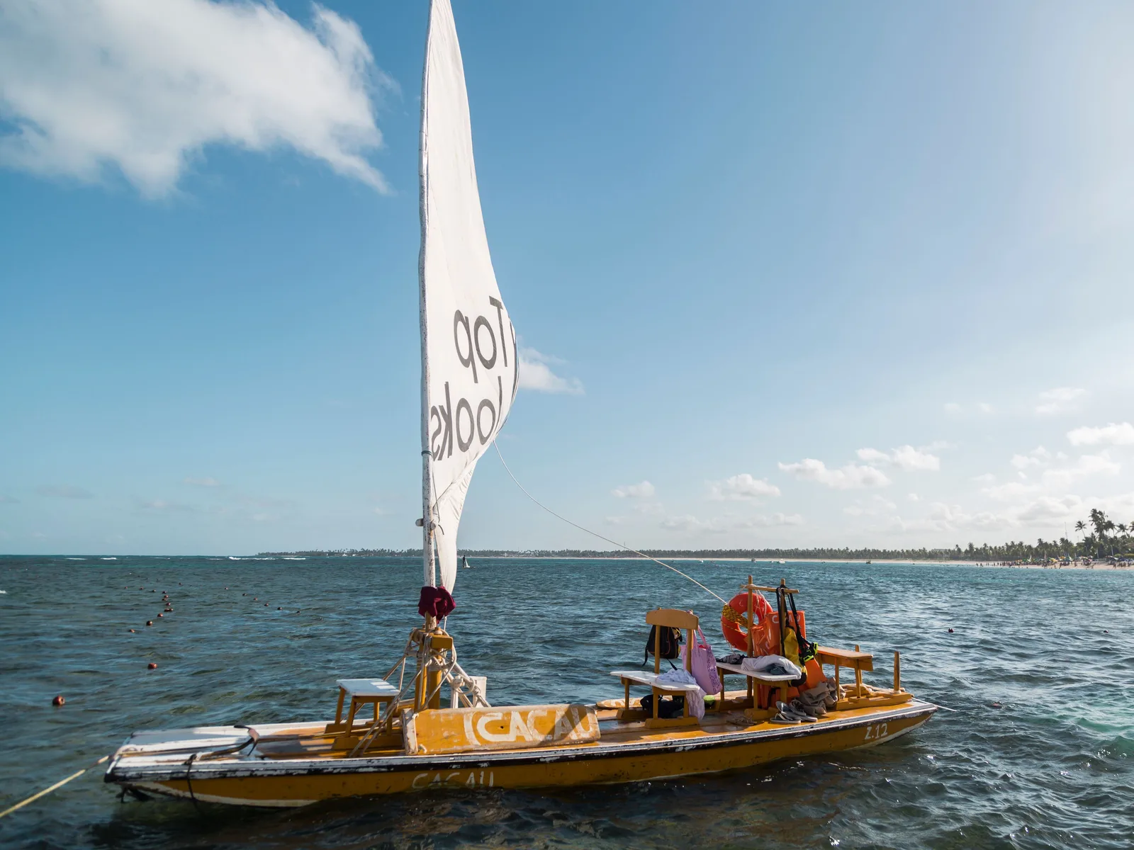 A jangada under a white sail on open green water off Porto de Galinhas with passengers aboard and the low green coastline behind