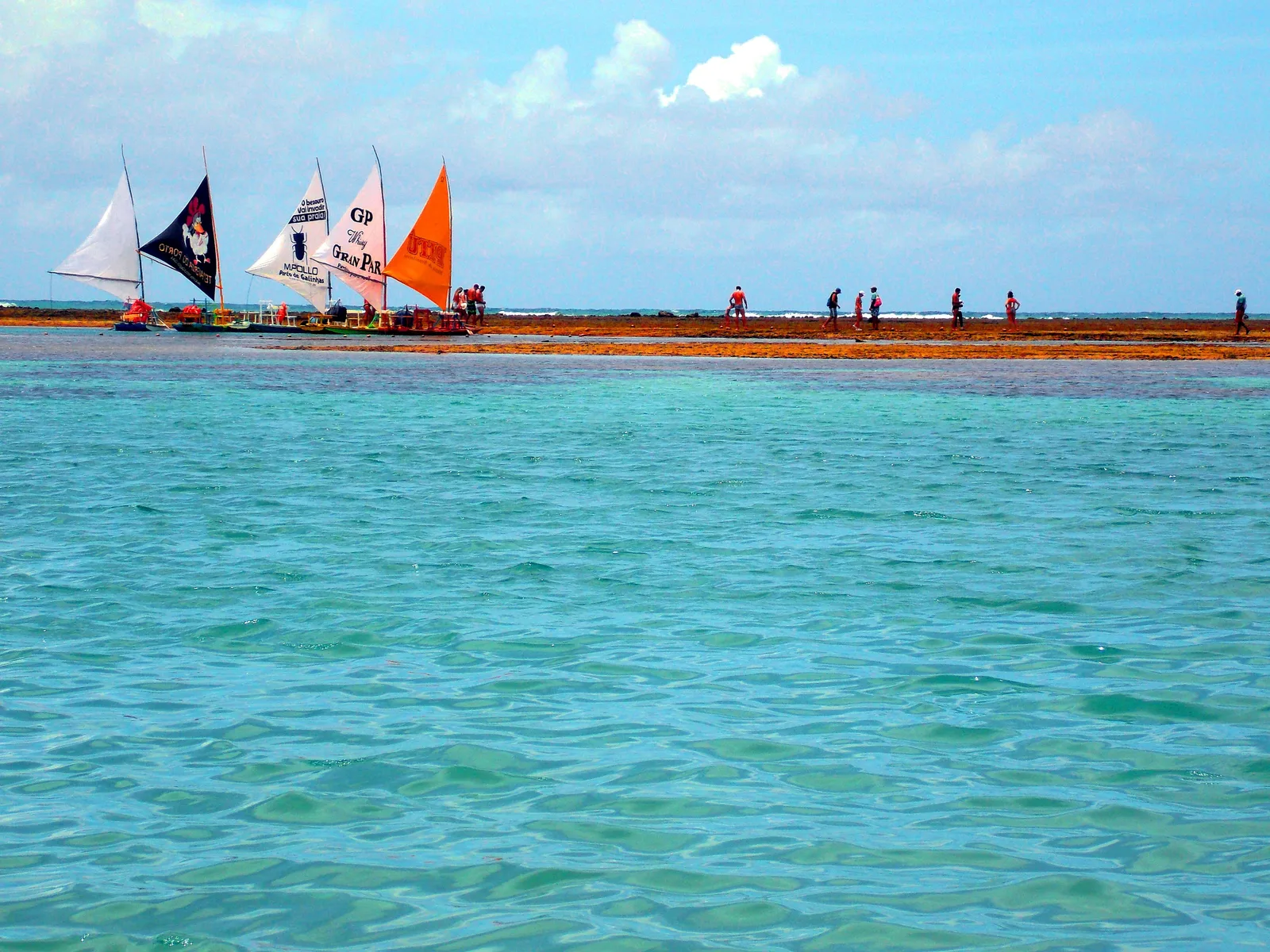 Jangadas with red, blue and orange sails moored in a line along the reef at Porto de Galinhas, people standing on the exposed rock beside them and a brilliant turquoise pool in the foreground