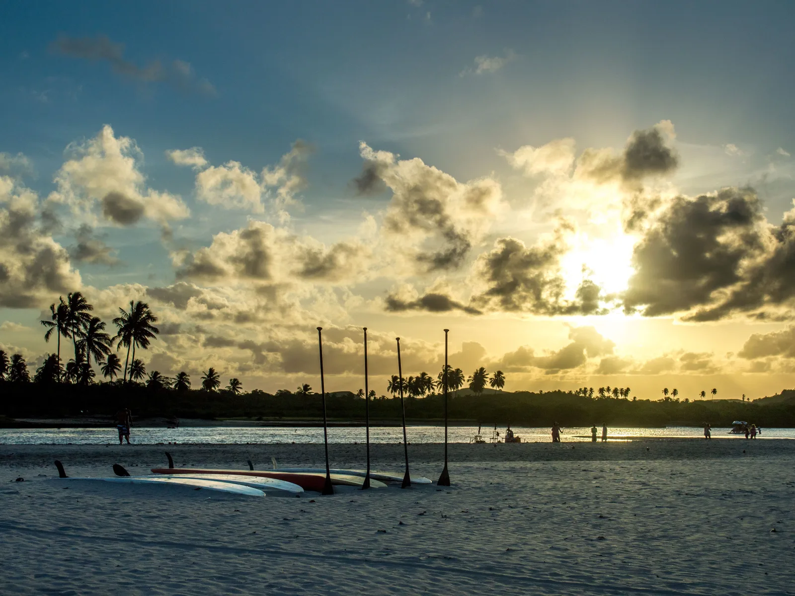Sunset over the shallow estuary at Pontal de Maracaípe, a row of wooden posts and a low jetty silhouetted against gold water with palms along the far bank