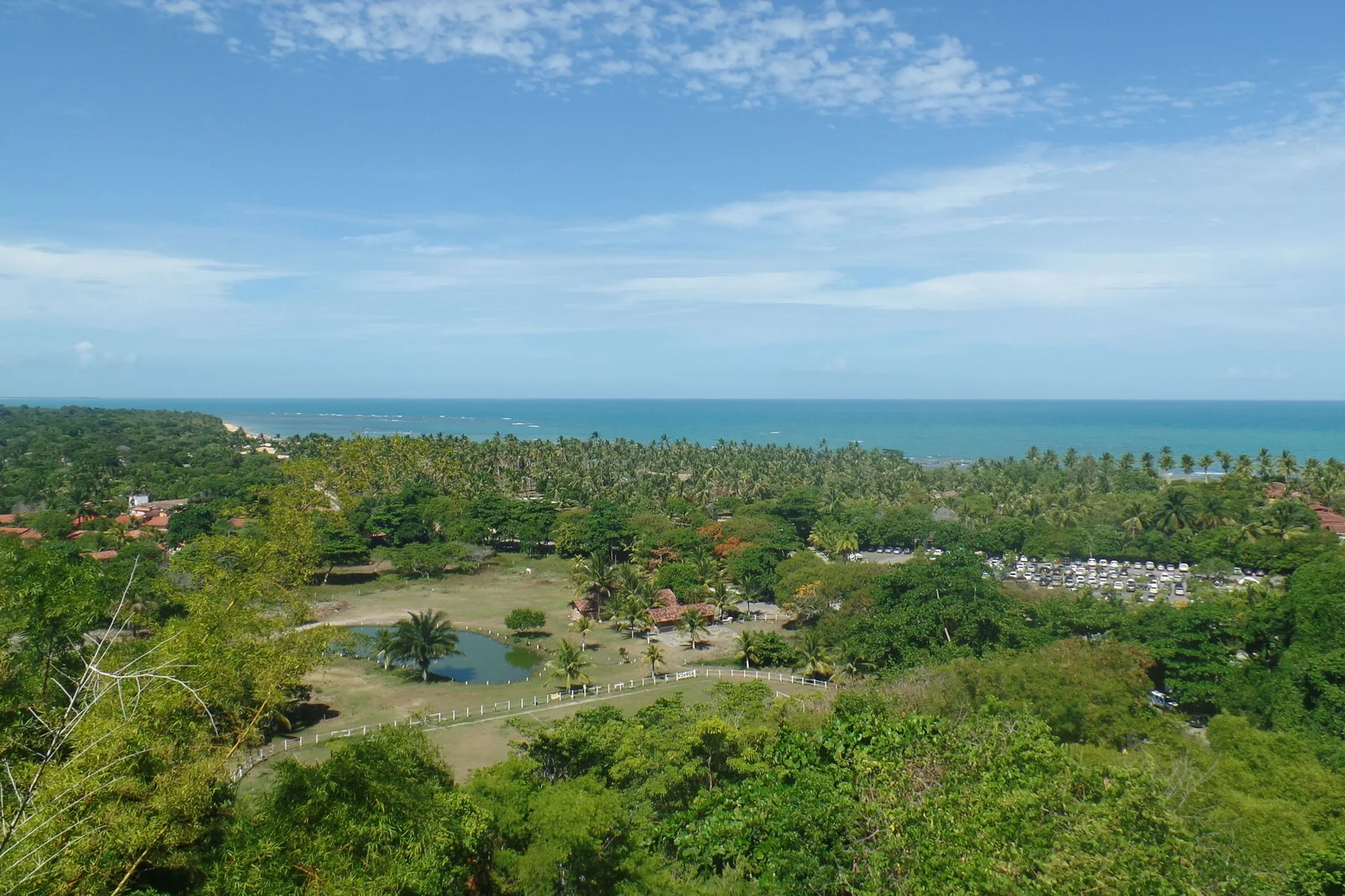 The view from the hill at Arraial d’Ajuda over dense green forest, a pond in a fenced paddock and a parking lot full of cars, with coconut palms and turquoise sea beyond