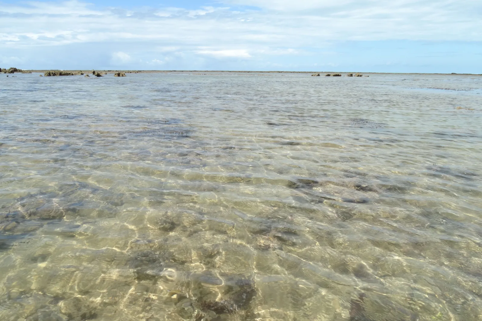 Clear, shallow, almost still water over sand and scattered rock at Praia do Espelho, with a dark line of reef showing along the horizon
