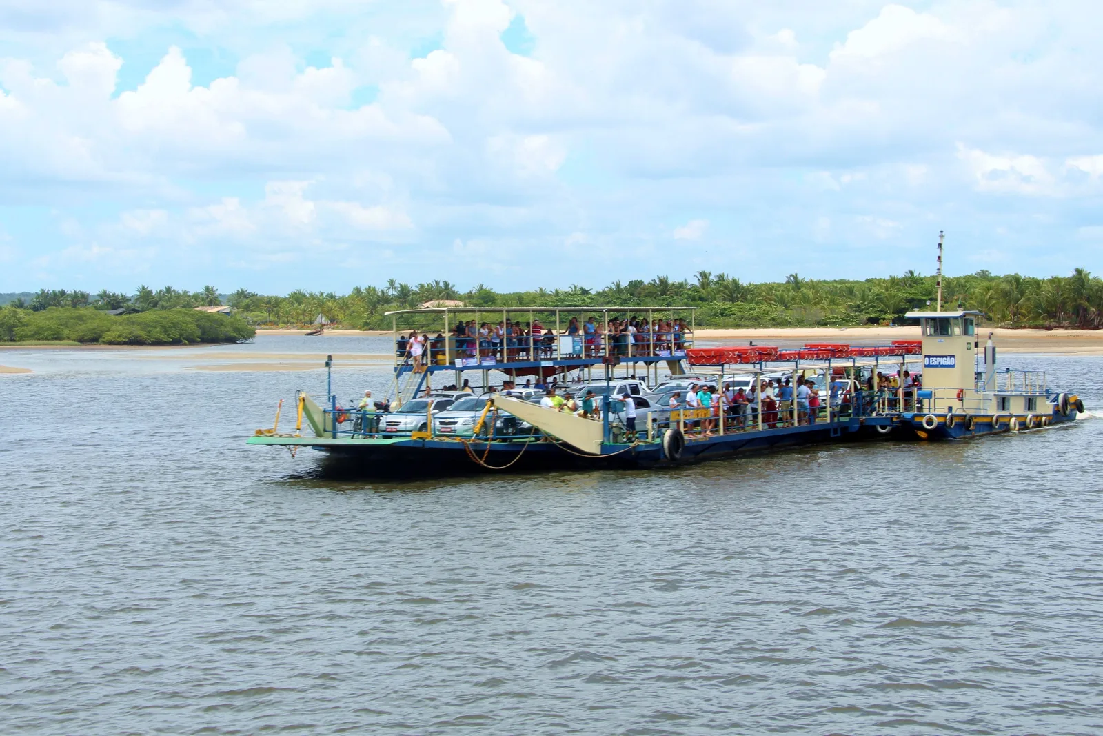 A flat vehicle ferry crossing the mouth of the Rio Buranhém, cars on its deck and foot passengers crowding a canopied upper deck, low green banks and a sandbar on the far side
