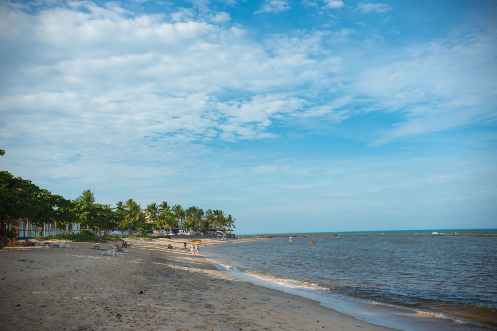The beach at Apaga Fogo: calm water along a wide sandy shore, loungers and a few people on the sand, and palms with low white buildings behind