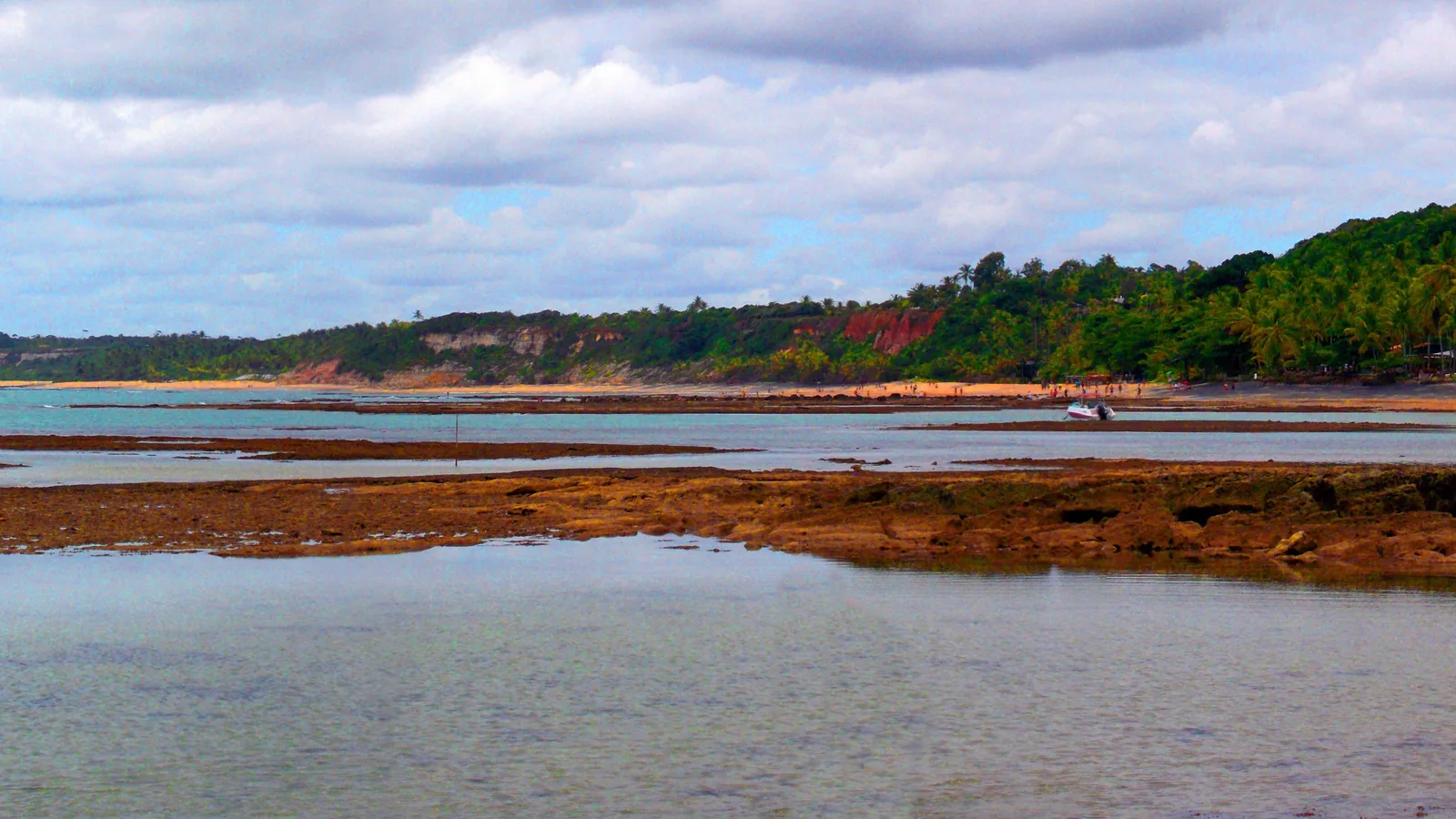 Low tide at Praia do Espelho: a flat of brown reef exposed across still, shallow water, with a line of low reddish cliffs and forest along the far shore under a cloudy sky